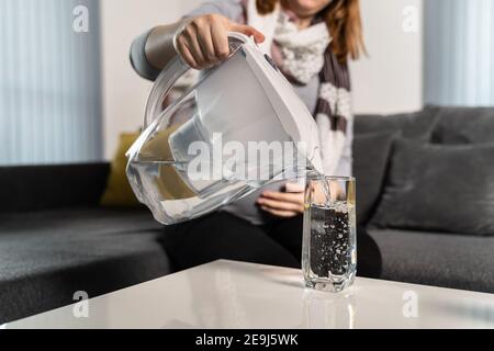 Donna sconosciuta che versa acqua filtrata in vetro sul tavolo dalla caraffa con filtro in plastica a casa durante il giorno - idratazione dell'acqua pulita concetto di vita sana Foto Stock