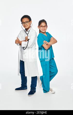 Carino bambini che indossano un uniforme medica con le braccia incrociate su sfondo bianco. Due bambini giocano al personale medico e scelgono una professione medica Foto Stock