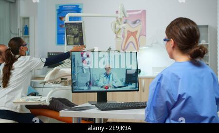 Infermiere discutere di consultazione con dentista senior medic su videochiamata in studio dentistico, mentre donna dottore sta lavorando con paziente in background. Medic di ascolto assistito sulla webcam Foto Stock