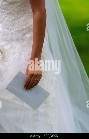 Mano della sposa che tiene una busta bianca da un ospite e anello di nozze alla sua mano. Il suo vestito da sposa bianco è sfondo. Foto Stock