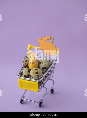 Preparandosi per la vacanza di Pasqua, ha macchiato uova di quaglia biologiche fresche e coniglietto di souvenir in un carrello del supermercato in miniatura su sfondo lilla. Selectiv Foto Stock