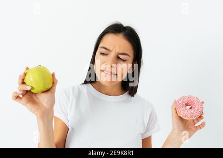 Giovane bella donna bruna sceglie tra cibo sano e dolci, guarda seriamente la mela che tiene la ciambella in altra mano. Foto Stock