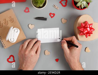 Qualcuno che scrive una carta vicino alla scatola del regalo e cuori sopra vista dall'alto del tavolo grigio Foto Stock