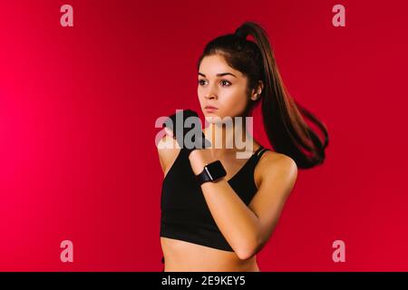 Ragazza runner concentrata in una parte superiore nera e orologio fitness su uno sfondo rosso guarda di lato. Spazio pubblicitario sul lato vuoto. Foto Stock