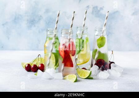 Tante bottiglie con rinfrescante limonata estiva con lime, fragole, ciliegie, cetrioli e ghiaccio su fondo grigio di cemento. Foto Stock