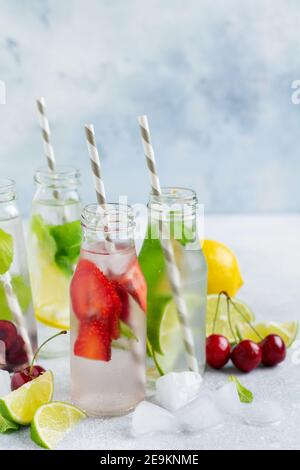 Tante bottiglie con rinfrescante limonata estiva con lime, fragole, ciliegie, cetrioli e ghiaccio su fondo grigio di cemento. Foto Stock