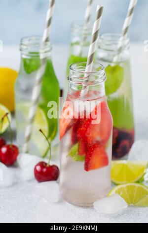 Tante bottiglie con rinfrescante limonata estiva con lime, fragole, ciliegie, cetrioli e ghiaccio su fondo grigio di cemento. Foto Stock