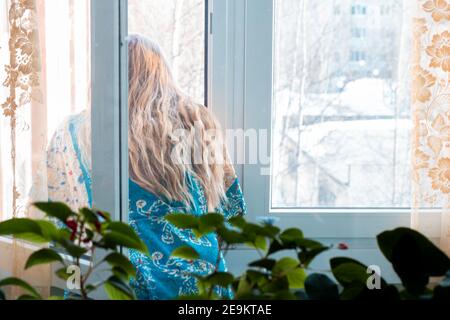 donna con lunghi capelli biondi si trova vicino a una finestra aperta e si affaccia sulla strada, casa quarantena auto-isolamento durante il periodo del coronavi Foto Stock