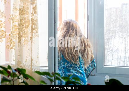 donna con lunghi capelli biondi si trova vicino a una finestra aperta e si affaccia sulla strada, casa quarantena auto-isolamento durante il periodo del coronavi Foto Stock