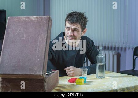 Salute psicologica. Un uomo si siede in cucina, beve alcolici e ascolta la musica su un gramofono Foto Stock