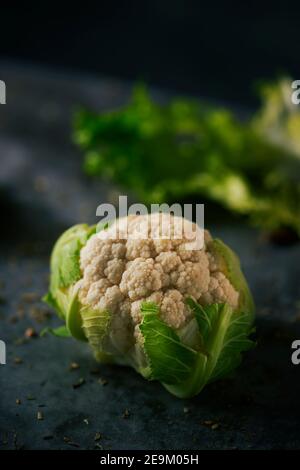 closeup di una testa cruda del cavolfiore del bambino posta su a. superficie in pietra scura cosparsa di semi e spezie diversi Foto Stock