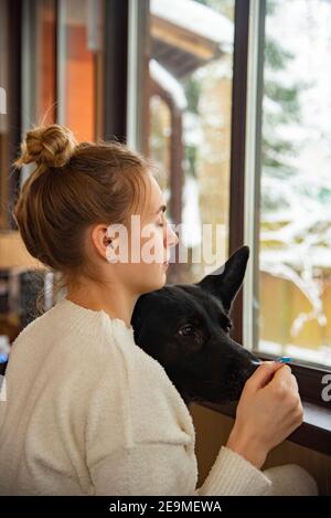 Una bella ragazza bionda in un vestito bianco casa e un cane pastore alla finestra con una vista sugli alberi innevati. Foto Stock