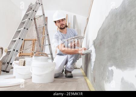 uomo addetto alla costruzione di intonaci al lavoro, prende l'intonaco dal secchio e lo mette sulla cazzuola per intonare la parete, indossa il casco all'interno del cantiere Foto Stock