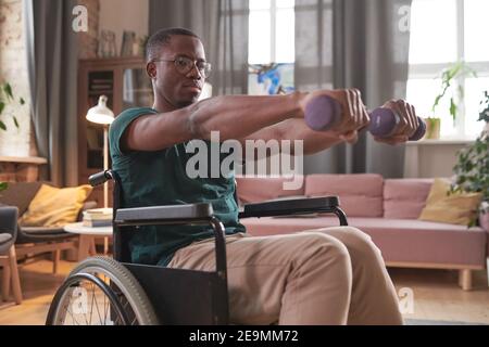 Uomo africano disabili seduto in sedia a rotelle e allenandosi con manubri a casa Foto Stock