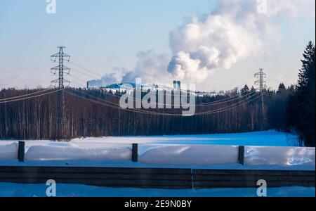 Helsinki / Finlandia - 5 FEBBRAIO 2021: Una linea elettrica con alti tralicci in piedi su un campo innevato. Due camini sullo sfondo. Foto Stock