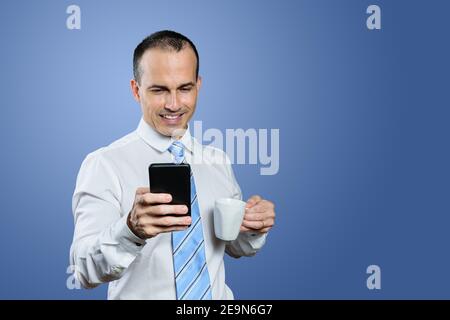 Sorridente uomo brasiliano maturo in abiti formali che tengono uno smartphone e una tazza. Sfondo blu. Foto Stock