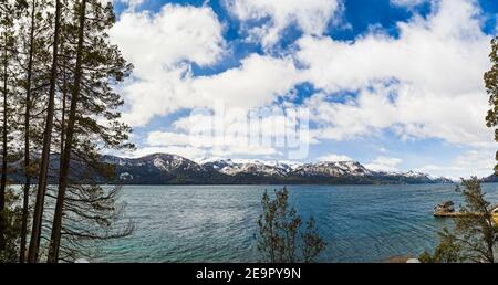 Splendida vista panoramica sul paesaggio con il lago Traful circondato da alberi alti, montagne innevate e molo sotto il cielo nuvoloso su una sorgente soleggiata Foto Stock