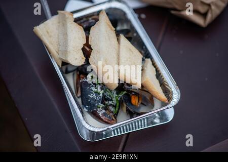 Sul tavolo è presente una lamina monouso contenente cozze cotte al fuoco in salsa all'aglio, cosparse di parmigiano e una baguette fresca croccante. Str Foto Stock