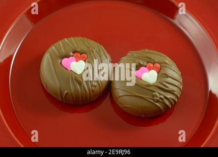 Due`s biscotti sandwich immersi al cioccolato di San Valentino con decorazioni a cuore su piastra rossa su sfondo rosa Foto Stock