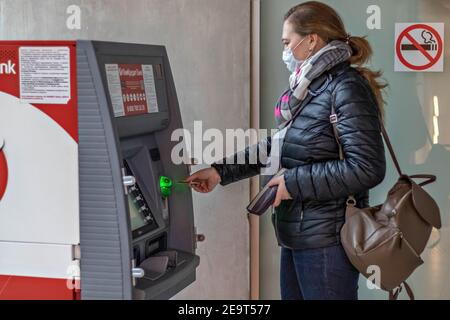 Una donna che indossa una maschera medica protettiva sul viso inserimento di una carta di credito e prelievo di contanti da un BANCOMAT Foto Stock