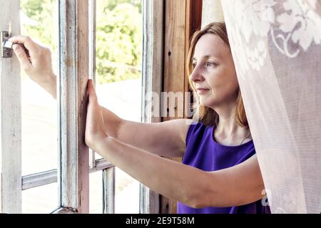 Giovane donna apre la finestra nella sua casa di campagna, guarda il giardino. Foto Stock