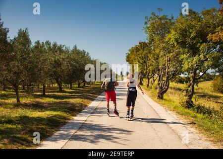 Vista posteriore di una coppia di amanti del pattinaggio su rollerblade sulla strada accanto al solco di olive a Strunjan, Slovenia Foto Stock