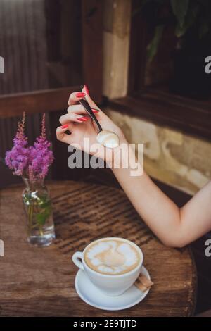 La mano di una donna tiene un cucchiaio con schiuma di latte da tazza di caffè su un tavolo di legno Foto Stock