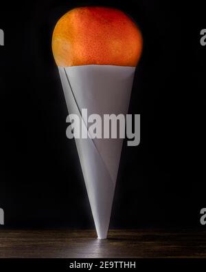 Un arancione è tenuto su da un cono di carta su una superficie di legno contro uno sfondo nero. Foto Stock