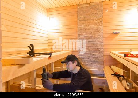 Costruzione di sauna, rifinitura. L'uomo sta avvitando una panca di legno al muro. Foto Stock