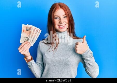 Giovane ragazza rossa della testa che tiene 10 libbre del regno unito banconote sorridenti felice e positivo, pollice su facendo eccellente e segno di approvazione Foto Stock