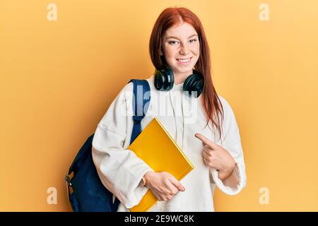 Giovane ragazza rossa della testa che indossa lo zaino dello studente e il libro di tenuta sorridente, con mano e dito Foto Stock