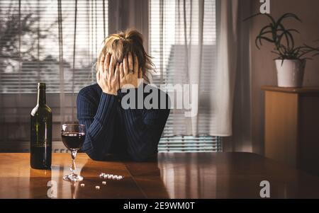 Donna depressa sta bevendo vino rosso e prendendo pillole a casa da sola. Testa nelle mani. Concetto di alcol e tossicodipendenza. Questioni sociali Foto Stock
