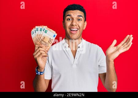 Giovane arabo che detiene 50 banconote in euro per celebrare il successo con buon sorriso e espressione vincente con mano sollevata Foto Stock