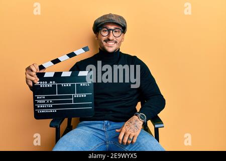 Bell'uomo con tatuaggi che tengono la clapboard del video film seduto sopra la sedia del direttore ha un aspetto positivo e felice in piedi e sorridente con un puzzore sicuro Foto Stock