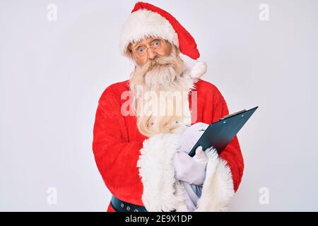 Vecchio uomo anziano con i capelli grigi e la barba lunga che indossa il costume di babbo natale che scrivono la lista indulgente e confusa espressione. Concetto di dubbio. Foto Stock