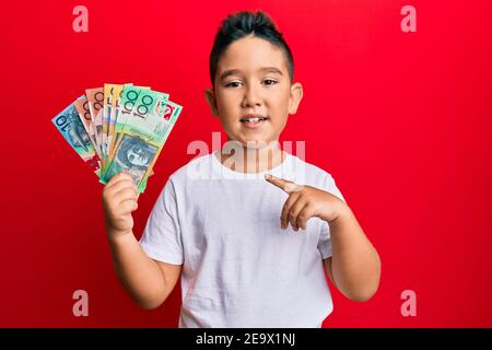 Ragazzino ispanico che tiene i dollari australiani sorridendo felice indicare con mano e dito Foto Stock