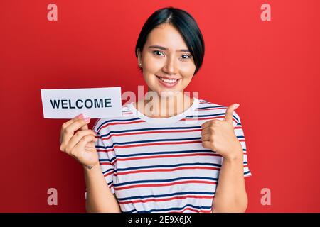 Bella giovane donna con capelli corti che tengono carta di benvenuto sorridente felice e positivo, pollice su facendo eccellente e segno di approvazione Foto Stock