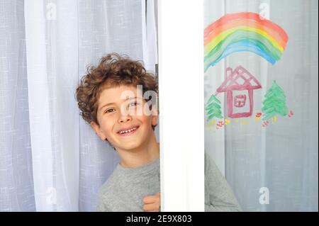 Carino ragazzino con un arcobaleno e una casa dipinta sulla finestra durante la quarantena a causa della pandemia del coronavirus. Foto Stock