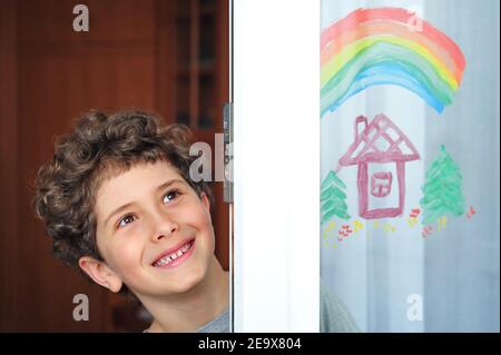 Carino ragazzino con un arcobaleno e una casa dipinta sulla finestra durante la quarantena a causa della pandemia del coronavirus. Foto Stock