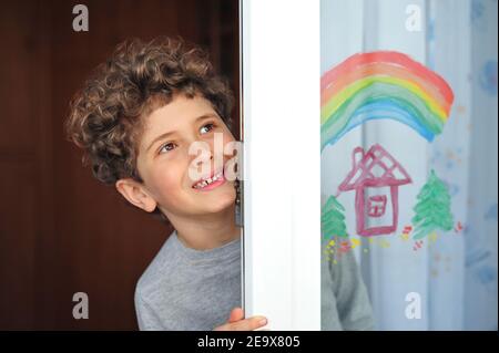 Carino ragazzino con un arcobaleno e una casa dipinta sulla finestra durante la quarantena a causa della pandemia del coronavirus. Foto Stock