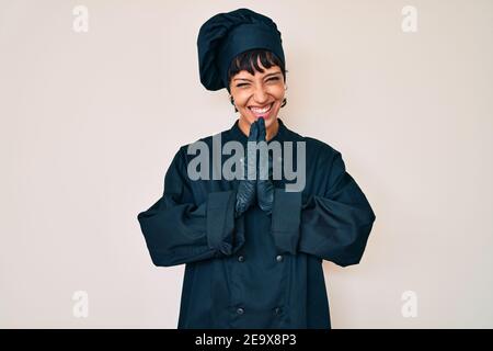 Bella donna brunettte che indossa una cuoca uniforme professionale che prega con le mani insieme chiedendo perdono sorridente fiducioso. Foto Stock