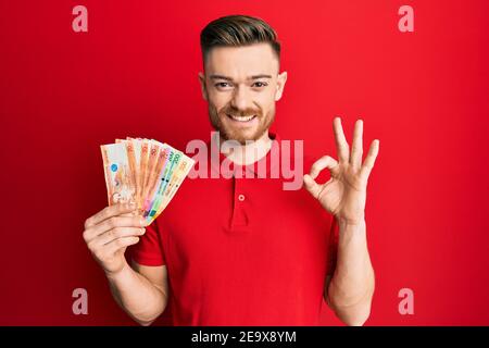 Giovane uomo rosso che tiene le banconote di peso filippino facendo segno ok con le dita, sorridente amichevole gesturing simbolo eccellente Foto Stock