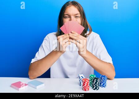 Bell'uomo caucasico con capelli lunghi giocando a poker che copre faccia con carte sorridenti con un sorriso felice e fresco sul viso. Mostrando denti. Foto Stock