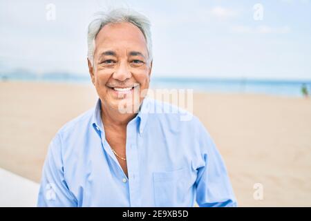 Hancosme uomo ispanico con capelli grigi sorridente felice in spiaggia, godendo vacanze in estate Foto Stock