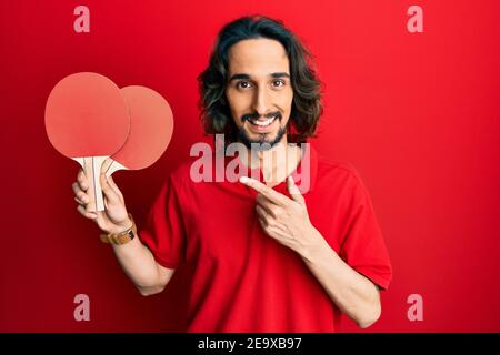 Giovane ispanico che tiene racchette da ping pong rosse sorridendo felice puntare con la mano e il dito Foto Stock