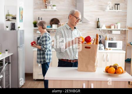 Coppia senior in arrivo dal supermercato con borsa della spesa e disimballaggio in cucina la mattina presto. Anziani pensionati che godono di vita, trascorrendo il tempo insieme aiutandosi a vicenda Foto Stock