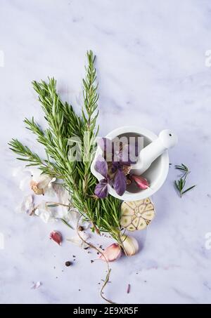 Dall'alto pestello e mortaio con fiori in fiore vicino all'aglio chiodi di garofano e rosmarino fresco su tavola di marmo Foto Stock