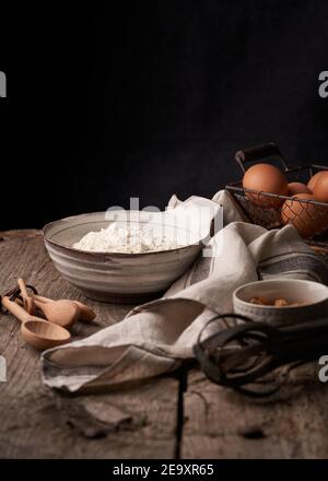 Un mucchio di uova nel cestello e una ciotola di farina messa su un tavolo di legno con vari cucchiai e frustate prima di cuocere Foto Stock