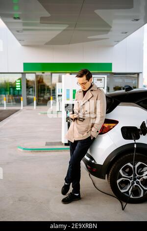 Imprenditore maschile in abito elegante in piedi alla stazione di benzina e. navigare sul telefono cellulare mentre si è in piedi vicino all'auto con la bocchetta carburante Foto Stock
