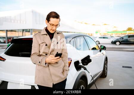 Imprenditore maschile in abito elegante in piedi alla stazione di benzina e. navigare sul telefono cellulare mentre si è in piedi vicino all'auto con la bocchetta carburante Foto Stock
